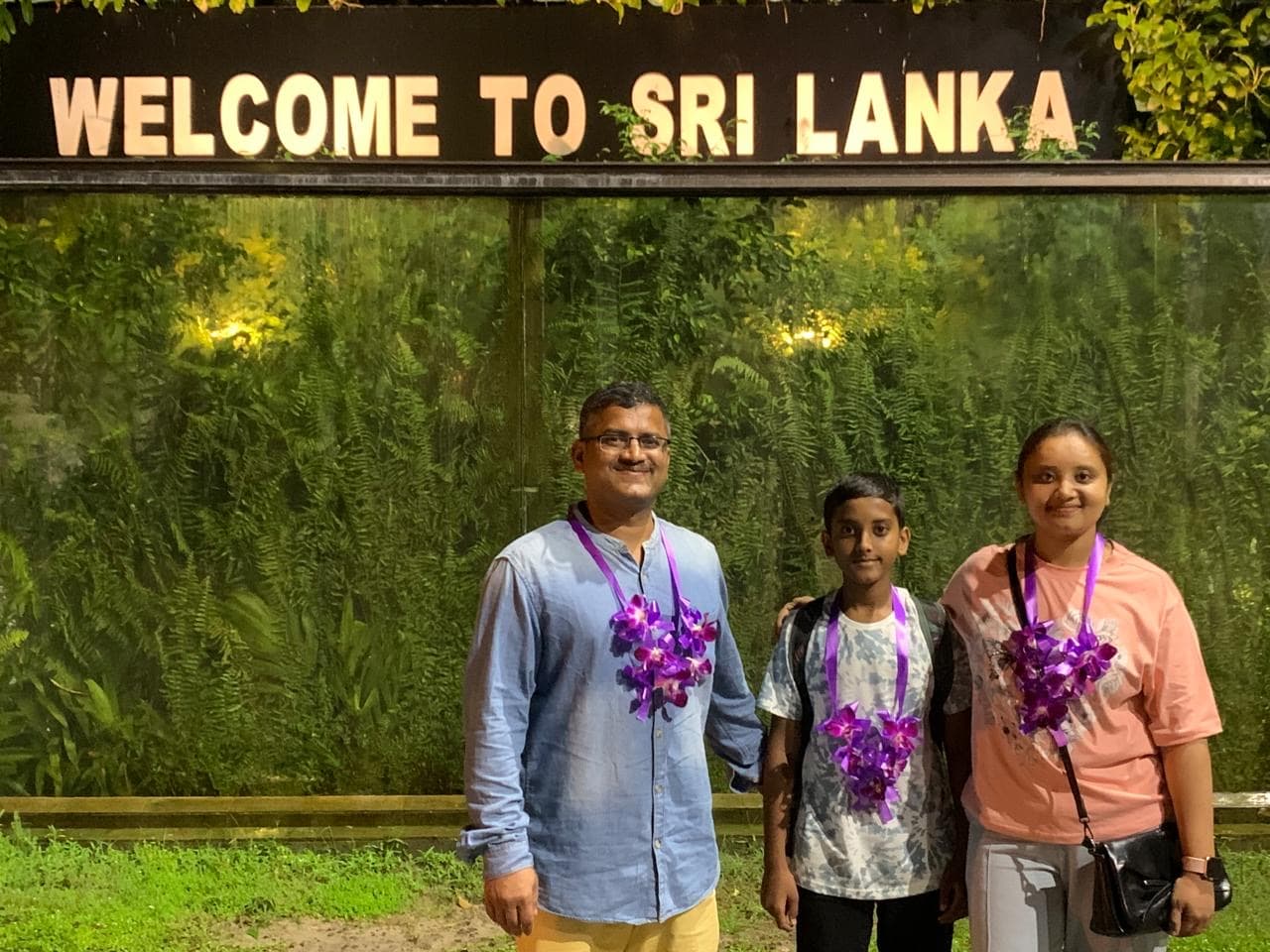 A family of three wearing purple orchid garlands beneath a Welcome to Sri Lanka sign