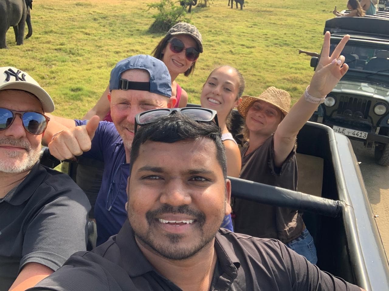 Guide and five guests in an open safari jeep with wild elephants in the grassland behind them