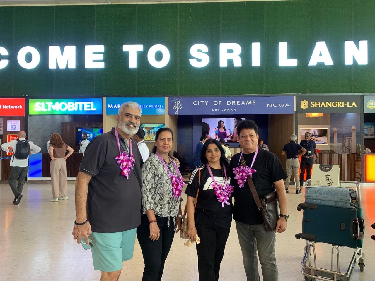 Four smiling guests wearing purple orchid garlands beneath a Welcome to Sri Lanka sign at the airport arrivals hall